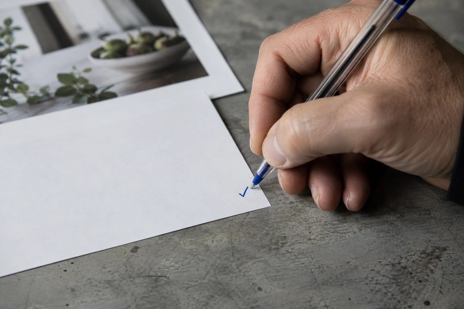 Eine Hand zeichnet mit einem Kugelschreiber ein blaues Häkchen auf eine freie Papierfläche.